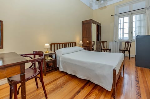 A bedroom with a white-sheeted bed, wooden furniture, hardwood floors, yellow walls, and natural light from windows with white curtains.